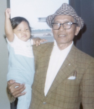 Photo of elderly man, Sylvestre Quitolis Carpio, holding an infant-aged Meda Higa. Carpio is using eyeglasses and wearing both a brown-white checkered hat and a dark khaki suit coat. Baby Meda is in a light blue jumper.