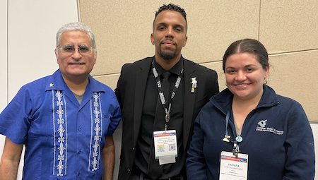 Group photo of speakers from SACNAS 2022 conference "Postdoc Fundamentals" session. Pictured from left to right Alberto Roca, Marcos Ramos-Benitez, and Yaihara Fortis Santiago standing in front of neutral tan background. Roca has short gray hair, wearing glasses, and in blue guayabera shirt with white vertical decorative pattern. Ramos-Benitez has black hair and goatee while wearing black coat and tie suit. Fortis Santiago has pulled-back dark brown hair and is wearing dark blue Sloan Kettering sweater.