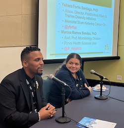 Photo of speakers from SACNAS 2022 conference &quot;Postdoc Fundamentals&quot; session. Pictured from left to right are Marcos Ramos-Benitez and Yaihara Fortis Santiago each sitting behind table-top microphones. Ramos-Benitez has black hair and goatee while wearing black coat and tie suit. Fortis Santiago has pulled-back dark brown hair and is wearing dark blue sweater. Behind them is the projected slide of their names in dark green font on a white background that also includes in red their Twitter accounts, @dr_mjramos and @yfortiss, respectively.